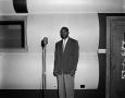 Photograph: [Photograph of unidentified man stands in front of microphone]