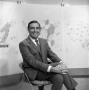Photograph: [Ron Godby sitting in front of weather maps]