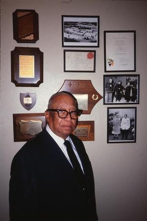 Primary view of object titled '[Dr. Thomas Tolbert stands in front of framed mementoes]'.
