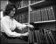 Photograph: [Woman standing beside bookshelf]