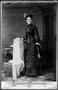 Photograph: [A woman standing beside a chair wearing a black dress and hat]