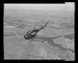 Photograph: [Photograph of a UH-1E Iroquois helicopter flying high over pastures]
