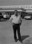 Photograph: [Man standing in front of a Ford dealership, 3]