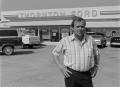Photograph: [Man standing in front of a Ford dealership, 2]