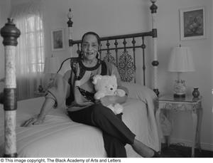 Black and white photograph of Dorothy Mayes Starks seated on the edge of a bed with ornate metal headboard. She holds a teddy bear in one arm.