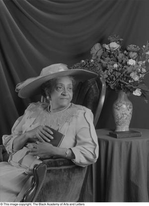 Black and white photograph of Mittie Dow Wise seated with a black draped fabric backdrop and bouquet behind her. She wears a large hat and holds a book.