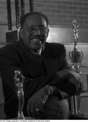 Black and white photograph of Marion Tawrice Jones seated, with his arms resting on a basketball in his lap, and basketball trophies surrounding him.