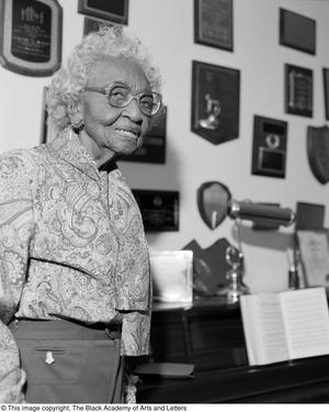 Black and white photograph of Marjorie Humber Jackson standing next to a piano, with many plaques lining the wall above.