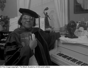 Fotografía en blanco y negro de la Dra. Francine Morrison de pie frente a un piano. Lleva una bata negra, un birrete de graduación de doctorado y sostiene un libro en el pecho.