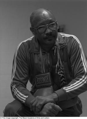 Black and white photograph of Curtis Cokes leaning his arm on one knee. He wears a track suite with stripes down the sleeves, and has a pair of boxing gloves draped around his neck.