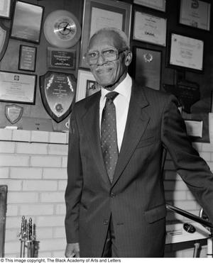 Fotografía en blanco y negro de John Clarence Phelps de pie frente a una pared llena de placas y documentos enmarcados. Lleva traje y corbata.