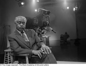 Black and white photograph of Jerome Thomas seated in a large empty sound stage, with a large television camera right behind him.