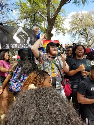 A crowd of people stand outdoors. One man is speaking into a microphone. A Black Lives Matter and rainbow pride flag are visible directly behind him.