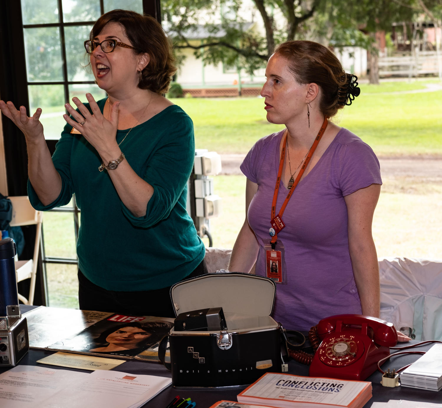 [Sixth Floor Museum booth], Photograph of the Sixth Floor Museum booth table at the DFW Archives Bazaar event inside of the Dallas Heritage Village pavilion. Curator Lindsey Richardson (green), and the Collection and Exhibits Manager, Stephanie Allen-Givens (purple), are behind the table and Richardson appears to be speaking to a visitor. There is a purse, telephone, LIFE Magazine, and several paper handouts on the table to look at., 