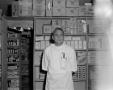 Photograph: [Druggist in front of shelf]