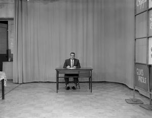 Primary view of object titled '[Man sitting at a desk on a tv set]'.
