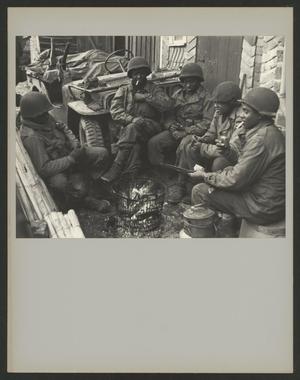 Black and white photograph of five young Black soldiers in military uniform with helmets. They sit circled around a small fire, two smoking cigarettes. A military jeep and wall can be seen directly behind them.