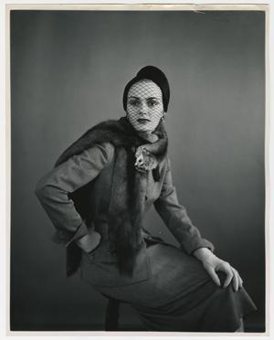 Black and white photo of a white woman sitting on a stool, body facing towards the right while eyes look just left of camera. She wears a suit with skirt, a dark hat with a mesh veil across her face, and a full animal fur stole around her shoulders. One hand sits on her knees while the other is on her hip.