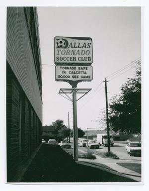 Un cartel que dice Club de fútbol del Dallas Tornado está en la parte superior, con otro letrero debajo que dice "Tornado seguro". Está junto a un edificio de ladrillos, con autos que se ven en el fondo.