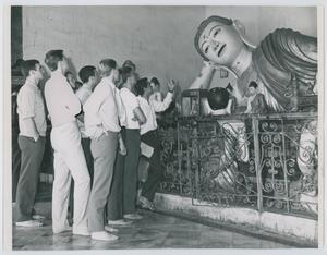 A group of men close together are seen looking at a giant buddhist statue, which is behind a fence.