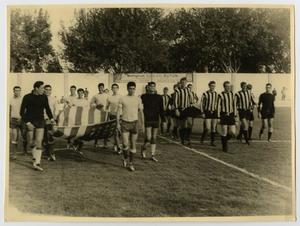Old photograph of men in soccer uniform, the group of men on the left walk with a flag extended out between them.