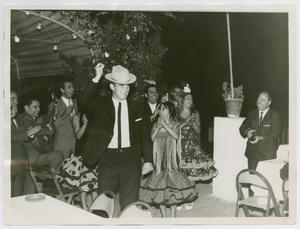 Foto en blanco y negro de un hombre con traje y corbata, y un sombrero de vaquero. Su brazo derecho está en el aire. A su alrededor hay hombres y mujeres riendo, las mujeres con vestidos.