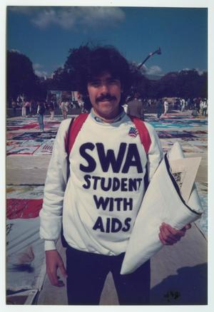 A man in a white sweater that says SWA, Stand With AIDS, holds a quilt in his hand while standing in front of a quilt display on the ground.