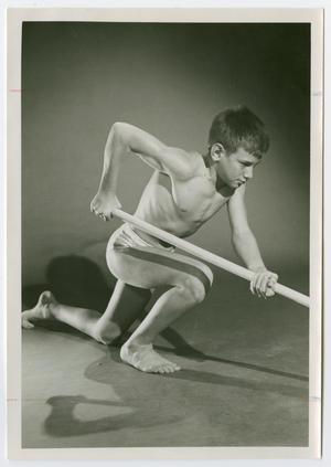 A young boy poses with his knees on the floor, wearing only underwear with a javelin extended out.