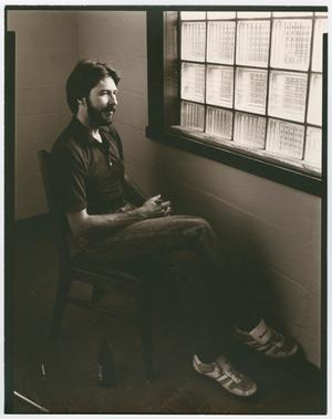 Man with a beard wears atheletic shoes, jeans and a black shirt. He sits at a chair facing a window. Sepia tone.