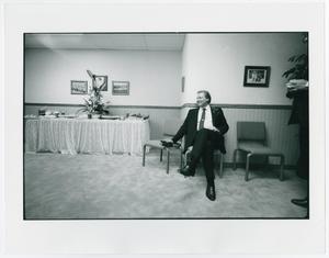 Un hombre con traje y corbata negros, sentado en una silla contra la pared. Él está en una habitación abierta, el lado izquierdo de la foto tiene una mesa cubierta con un mantel con una planta encima.