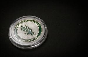 A silver coin with a green symbol of the eagle in the middle. Around it in a circle is a green line with words University of North Texas on it, and the date September 10, 2011 on it.