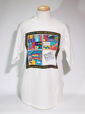 A white t-shirt with a square, quilt-like design on it. It is framed by a black line that says The NAMES Project. The middle has four different sections of design.