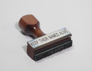 An ink stamper with a wooden handle. On it are the words Keep Their Names Alive.