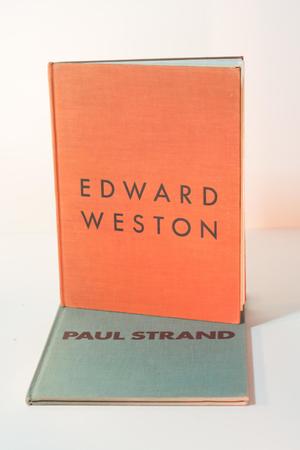 An orange book with the words Edward Weston on it in the middle. There is also a light blue book with the words Paul Strand in the middle.