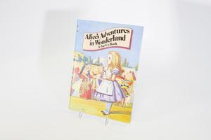 Una cubierta de libro que contiene una ilustración de Alicia de Alicia en el País de las Maravillas mirando hacia la izquierda. Se ven otros personajes detrás de ella y un cielo azul brillante por encima de ellos.