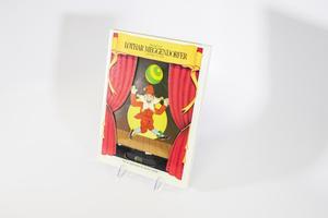 Una portada de libro con una ilustración en forma de marioneta en la parte delantera de una marioneta en  el centro de un escenario con los laterales mostrando cortinas rojas colgadas a los  laterales. La marioneta está de pie frente a una luna amarilla.