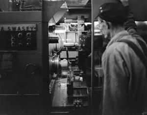 Black and white photo of an older man, with his back to the camera, looking into some dimly lit machinery. A panel of buttons is visible on the left side of the machine, while at center what looks to be a lathe is visible with water spraying all around.
