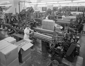 Black and white photo of rows of long machines producing what looks like paper items. Woman in white dress uniforms work around the machines, with stacks of large cardboard boxes sitting at the end of each machine.