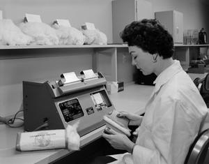 Black and white photo of a woman wearing a white lab coat sitting at a table that runs along the wall. A small machine with some sort of gauge on the face sits on the table in front of her. She holds two small devices in her hands with a piece of cotton sandwiched between. On the shelf in front of her, four large clumps of cotton sit with paper labels sticking out of their tops.