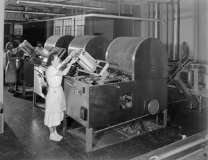 Foto en blanco y negro de dos mujeres delante de una fila de tres grandes máquinas. Las mujeres están apilando artículos planos en las máquinas. Llevan uniformes blancos.