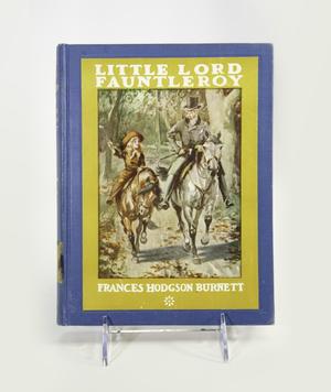 Photograph of the cover of the book Little Lord Fauntleroy, with the title in white text at the top, and author name at bottom. A rectangular illustration is at the center showing a young boy with long blond hair riding a small brown horse, and an older man to the right wearing a top hat and suit, riding a larger gray horse, on a path. The illustration is surrounded by a section of yellow, where the text sits, and the yellow is surrounded by blue.