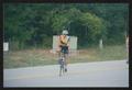 Photograph: [Cyclist in a red, blue and yellow jersey biking into a campground: L…