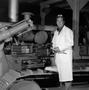 Photograph: [Man in a white coat posing by a large machine]
