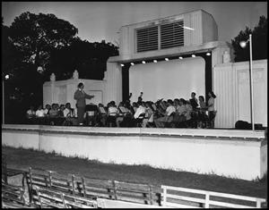 Foto en blanco y negro de una banda sentada en un escenario y el director frente a ellos.