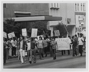 Foto en blanco y negro de personas con pancartas caminando a un lado del edificio. Una pancarta dice La hermandad es poderosa, otra dice Alguien a quien amas es gay.
