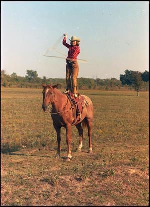 Woman in red shirt, brown pants and a cowboy hat stands on top of a brown horse. She swings a rope around her. They are on a field of grass.
