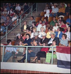 View of part of crowd sitting in seats. A metal rails is in front of the first row. The rows of seats go up, a rail in the middle of two sections and stairs going up.