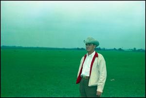 Man in a white jacket with a red collar wears a cowboy hat. He is standing outside with a green, even field behind him. The sky seems to be cloudy.