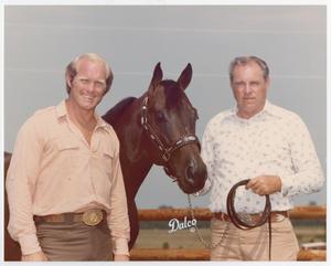 2 men stand stand with a horses head in between them. The man on the right holds the horses reigns in his left hand