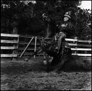 Black and white photo of a man in a cowboy hat riding a horse. The horse has its front legs in the air and dust is being kicked around him. Behind them is a wooden fence.